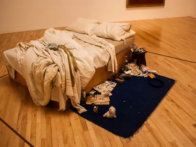 In this image, we can see a bed contains pillows and bed sheets. There is a carpet in the middle of...