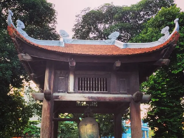 In the image there is a huge bell under a shelter and around that bell there are many plants and...
