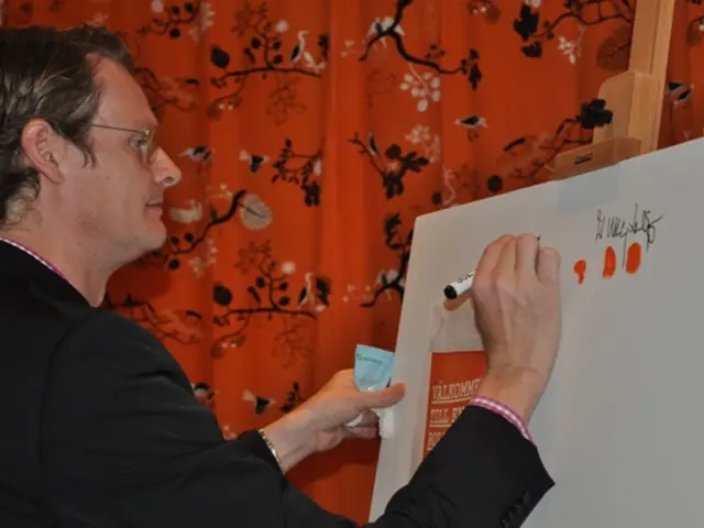In the foreground of the picture there is a person in black jacket writing on a whiteboard. On the...
