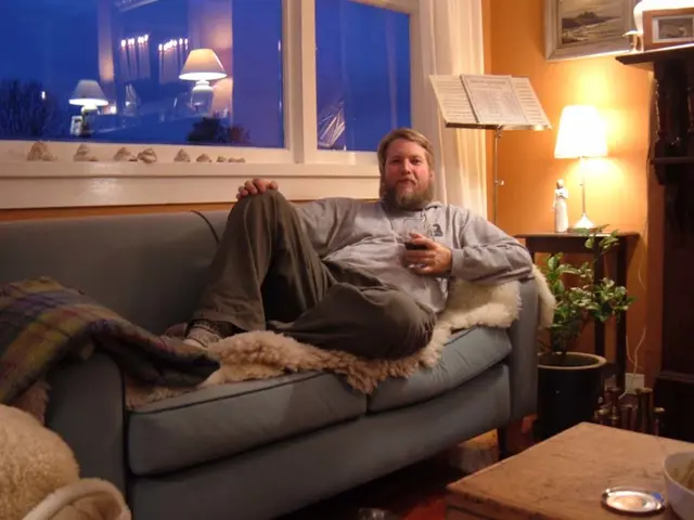 In this image the man is sitting on the couch. On the table there is a bowl and a plate. There is a...