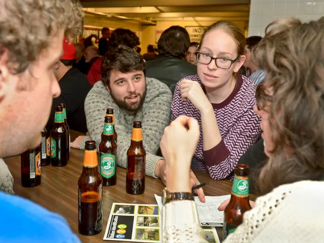 In the image there are several people stood on either sides of table with beer bottles on it. This...